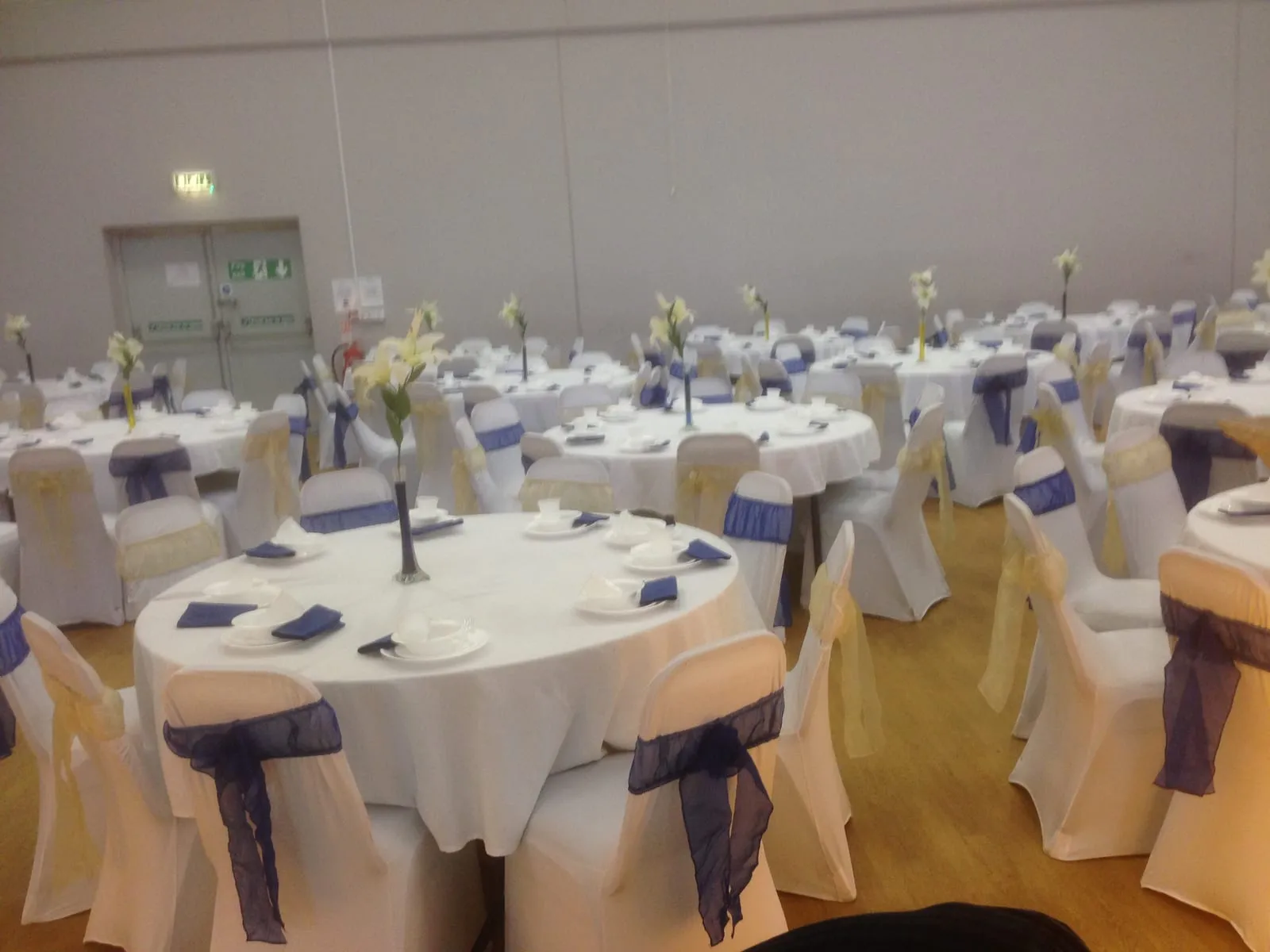 A large banquet across the whole hall: round tables with white cloths, navy and gold chair sashes and lily centrepieces.