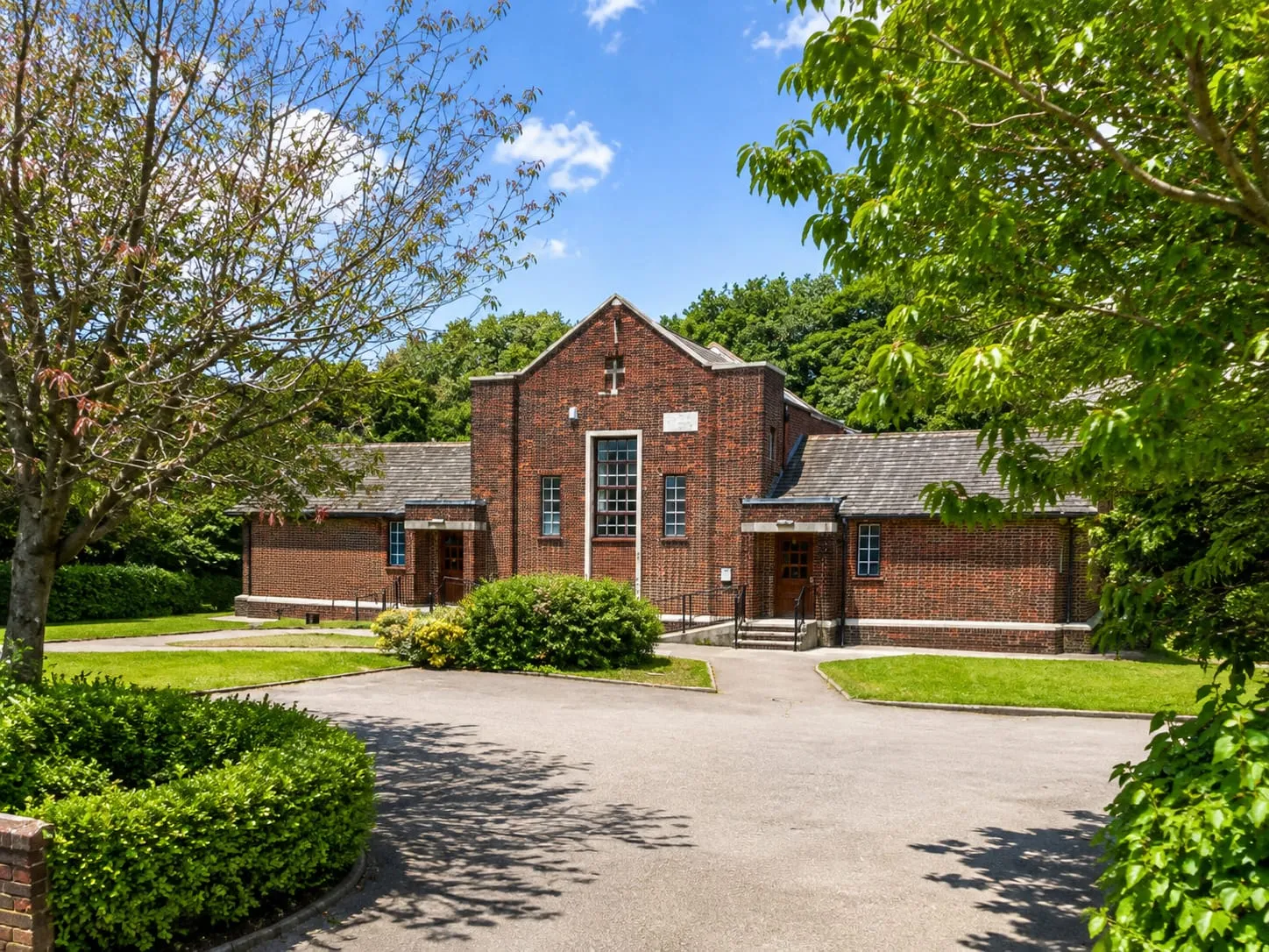 The original Brackenhall Community Centre on Longhill Road: a single-storey red brick building with a slate roof and a tall gabled entrance, set back from the road behind trees and a paved forecourt.