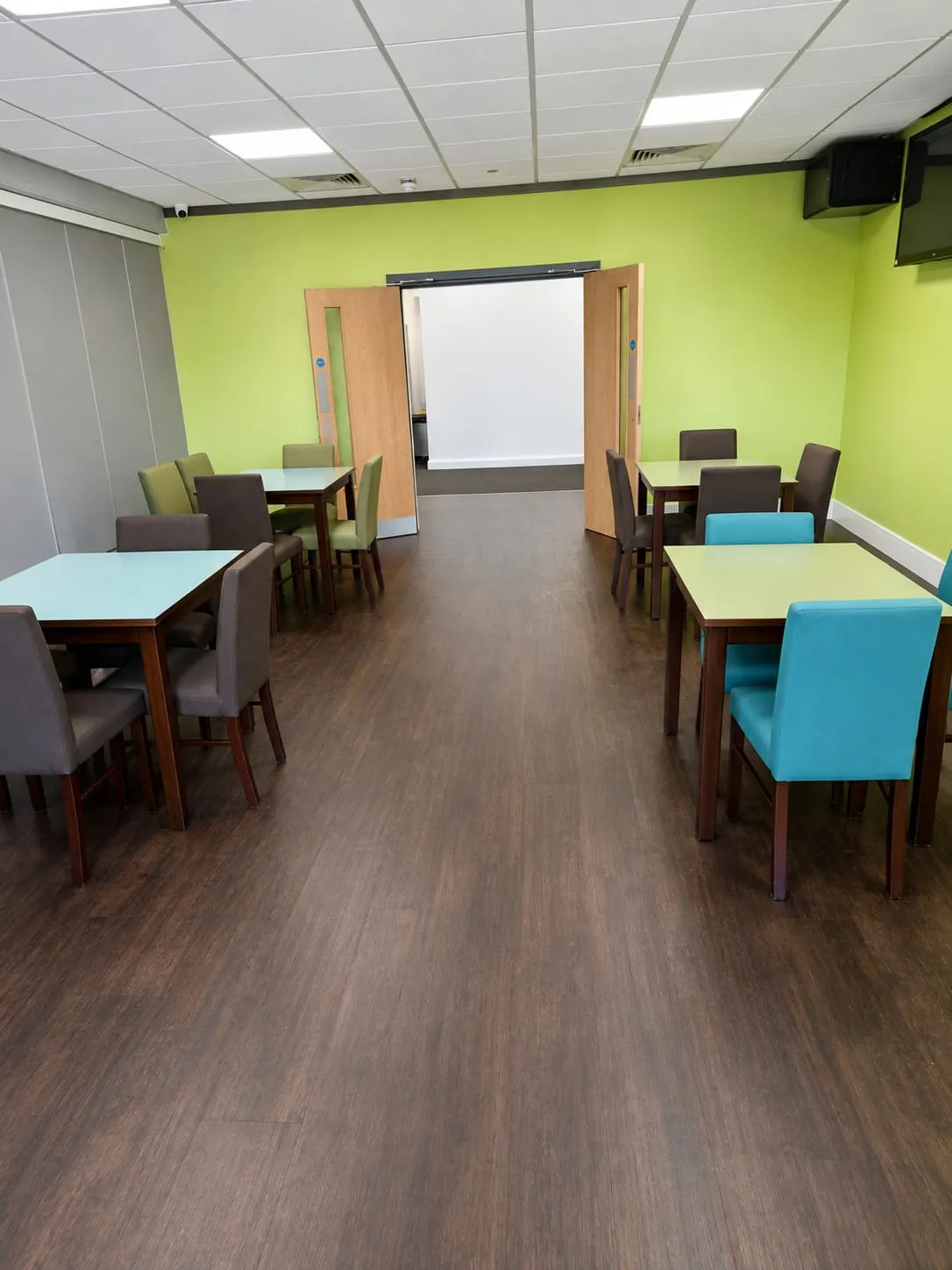 The canteen: square tables with pale green and blue tops and upholstered chairs, set out on a dark wood floor against a lime green wall, with wide double doors at the far end.