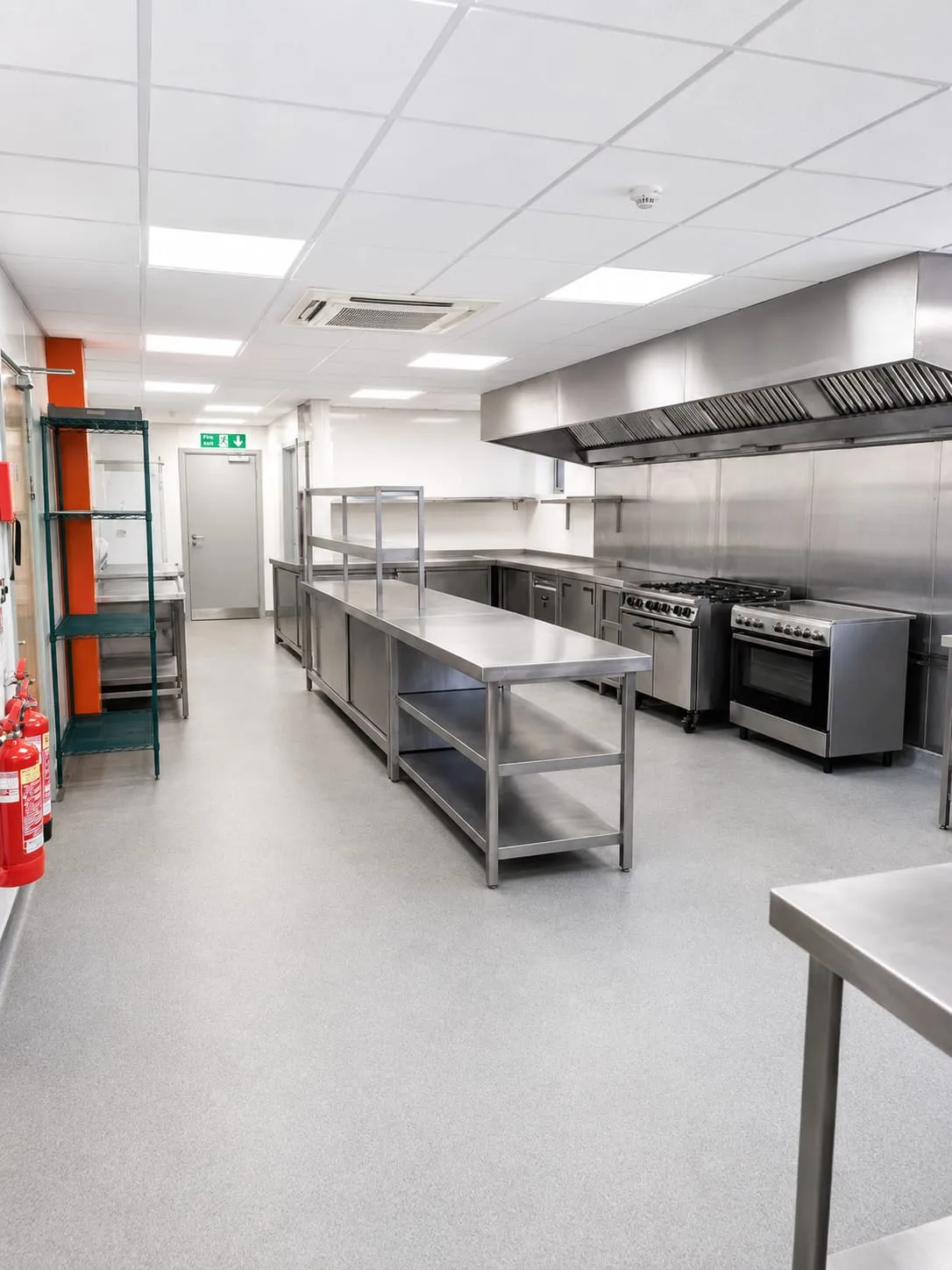 The commercial kitchen: a long stainless steel preparation table down the middle, a gas range and oven under an extraction canopy, and steel shelving along the wall.