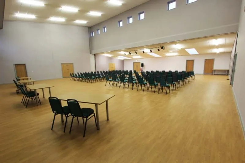 The main hall with the partition wall open, set with rows of chairs facing a stage area and a few tables to one side.