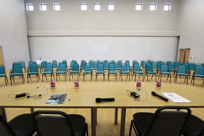 A long top table with microphones in the foreground, facing rows of teal chairs across the hall.