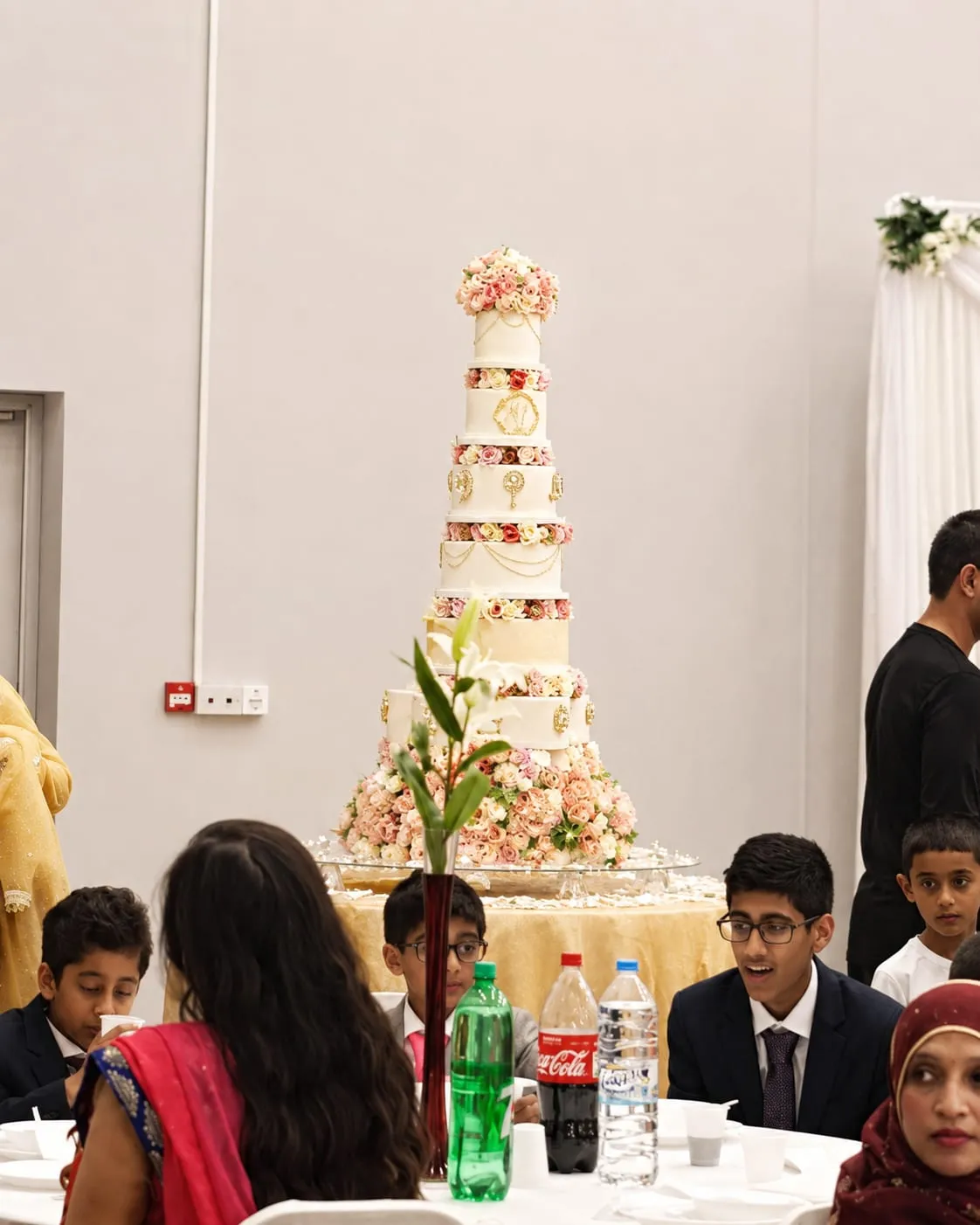 A tall multi-tier wedding cake decorated with roses on a table at the edge of the Whole Hall, with guests seated nearby.