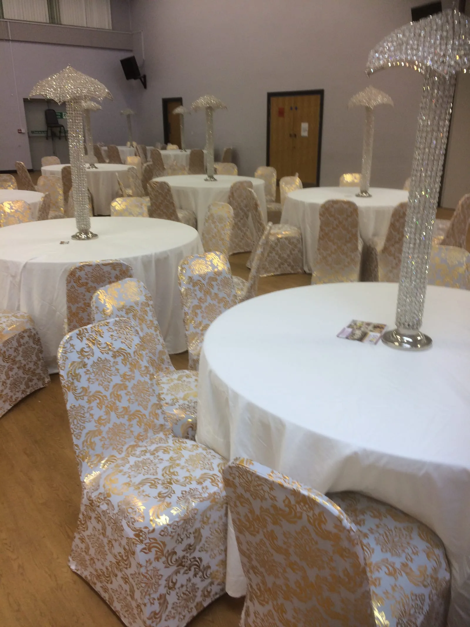 Round tables with white cloths, gold damask chair covers and tall crystal centrepieces, dressed for a wedding.