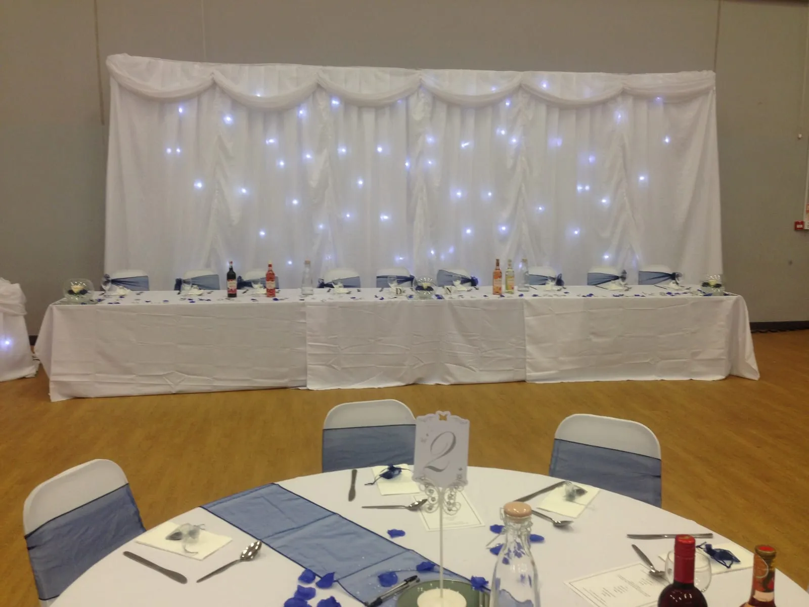 A long top table in front of a white starlight curtain backdrop, with blue chair sashes and blue table runners.