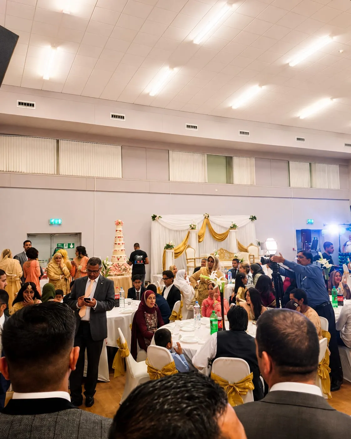 A wedding reception filling the Whole Hall, with round tables in gold-sashed chair covers, a decorated stage backdrop and guests seated and mingling.